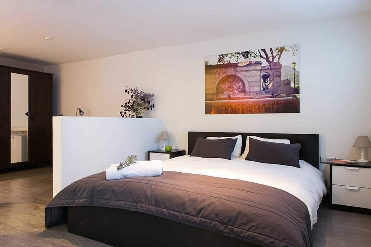 A modern hotel room with a large bed, a nightstand, a wall-mounted painting, and a door to the left.