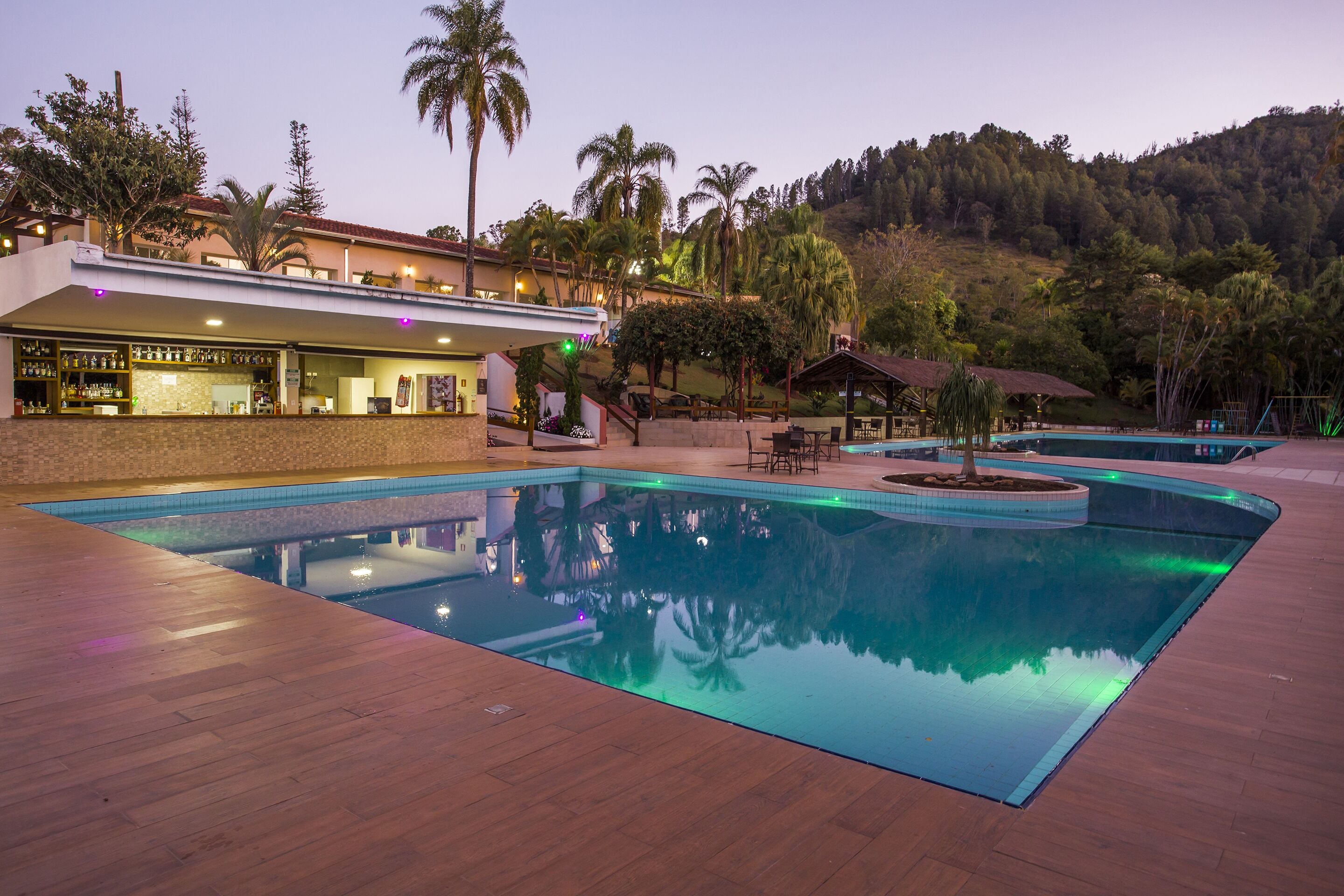 A pool area with a bar and outdoor seating, surrounded by palm trees and a hilly landscape.