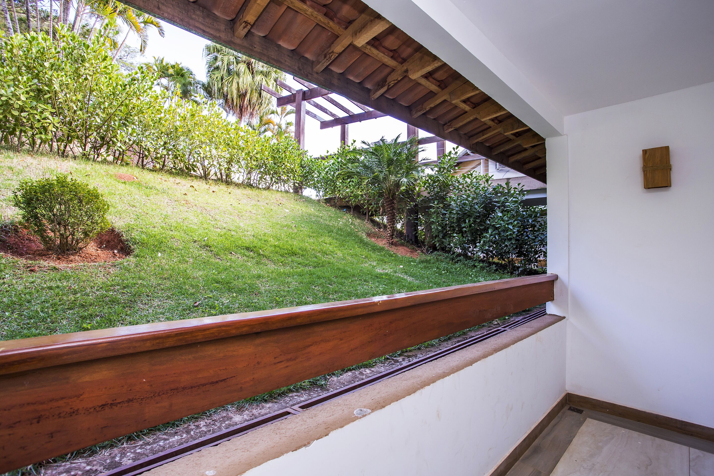 Un balcon doté d’une balustrade en bois, donnant sur un jardin bien entretenu où poussent différentes espèces de plantes, ainsi qu’un palmier.