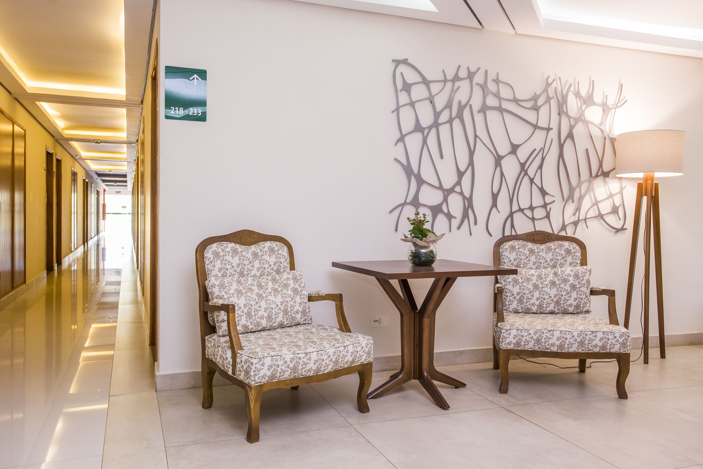 Um corredor com duas poltronas estofadas, uma mesa, uma obra de arte na parede e uma placa de sinalização.