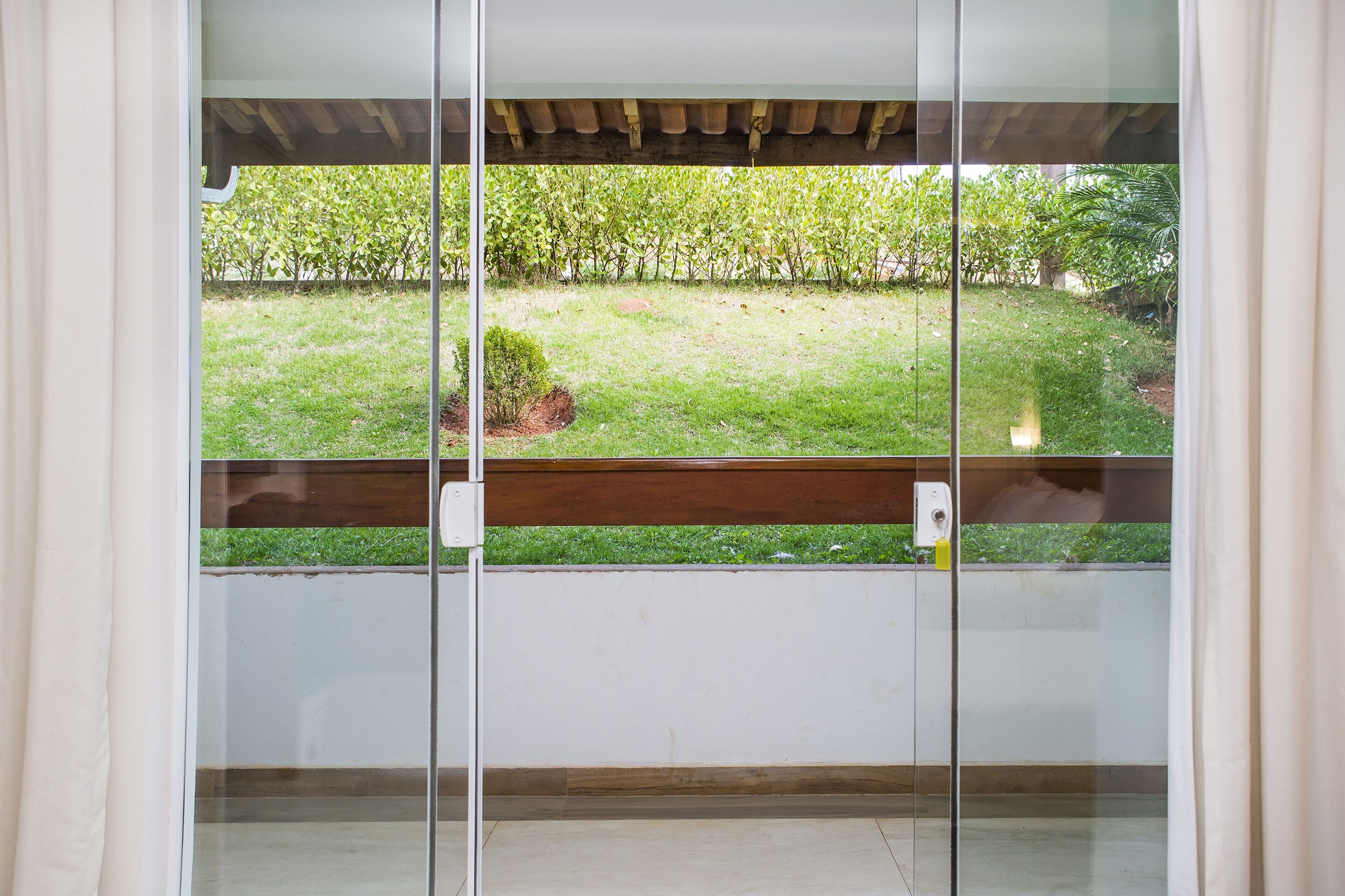 A view through a glass door to a balcony with a wooden railing, a small garden area with grass and shrubs, and a white wall in the background.