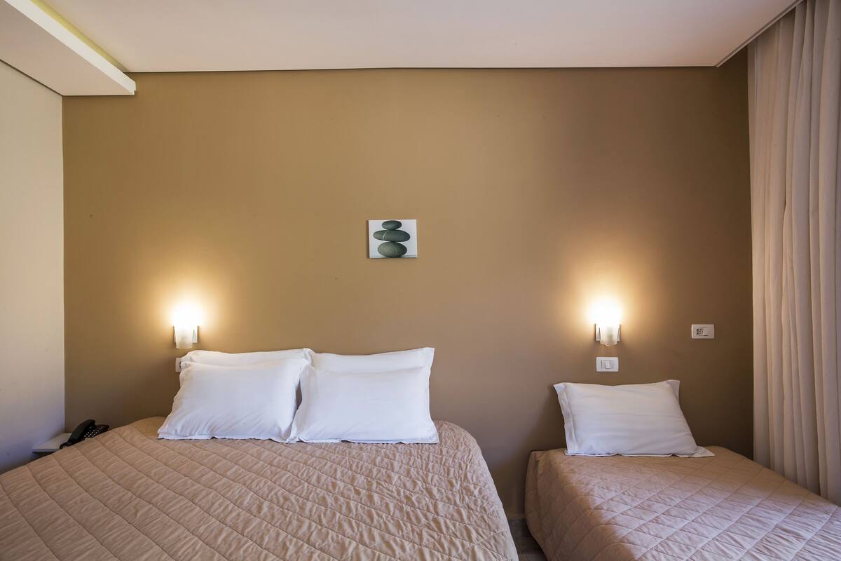 Habitación de hotel con dos camas, cada una con ropa blanca y colcha acolchada color beige. Hay lámparas de pared a ambos lados de las camas, y un pequeño cuadro está colgado en la pared entre ellas.