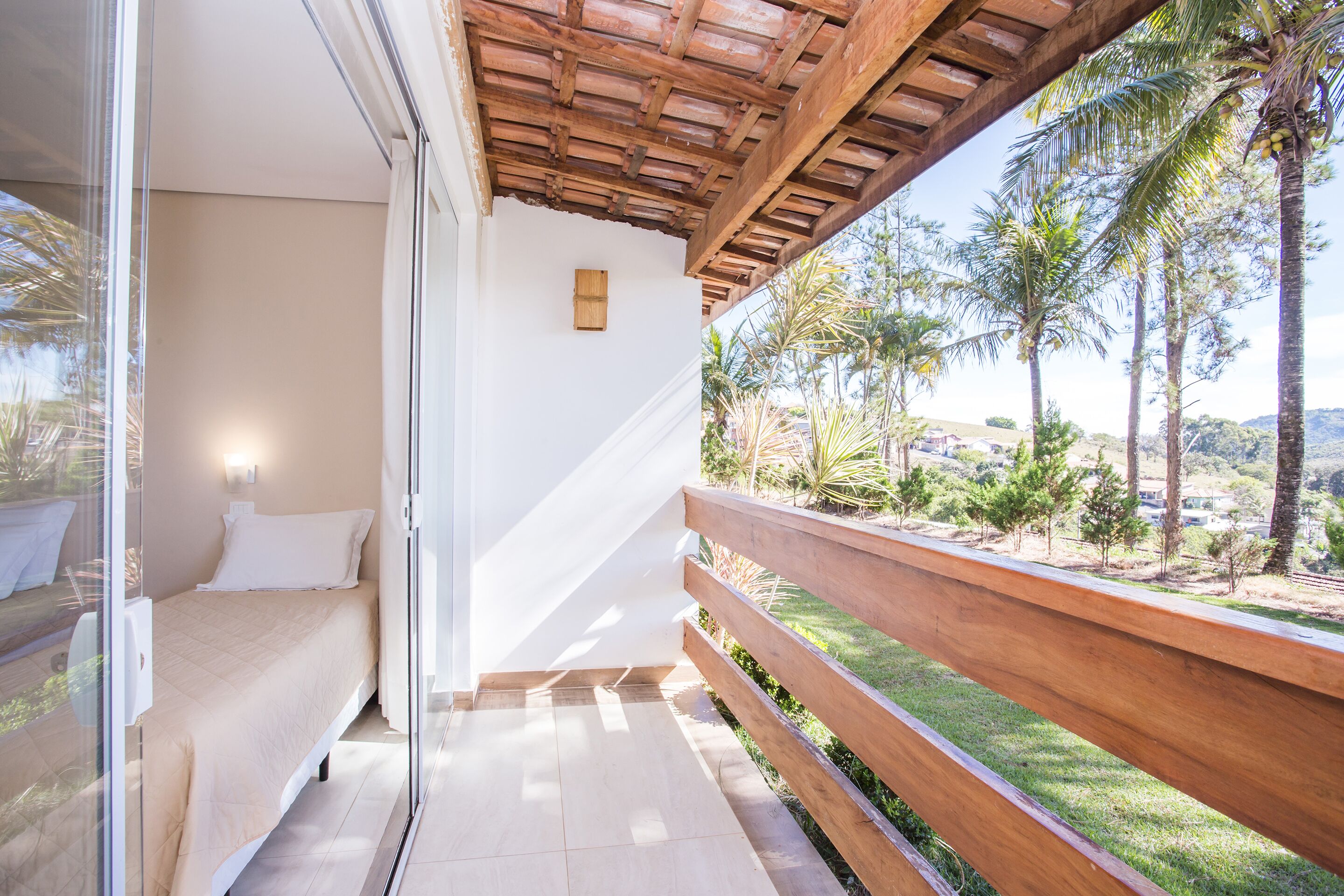 A balcony with a wooden railing, a bed with white bedding, and a view of palm trees and greenery.