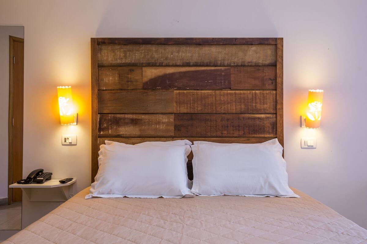 Una cama bien hecha con dos almohadas, un cabecero de madera, lámparas de noche y una mesita con un teléfono y un mando a distancia.