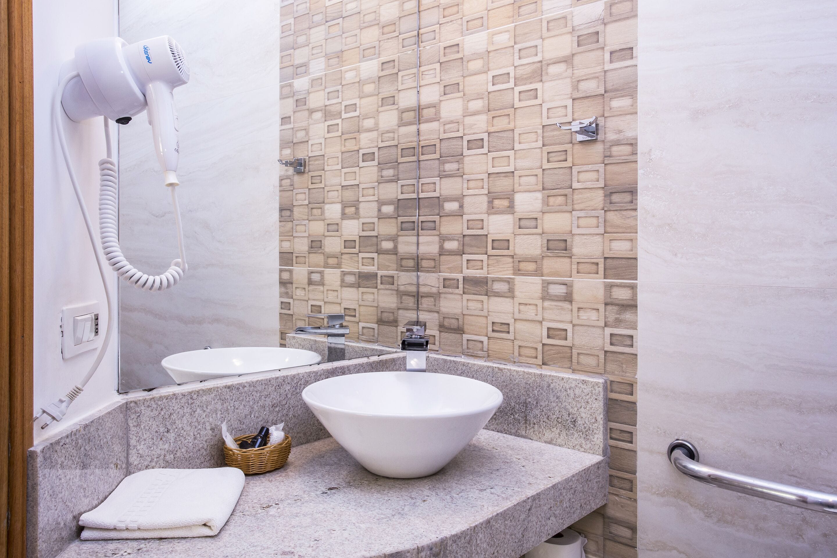 Un baño con un lavamanos blanco, un espejo grande, un secador de pelo montado en la pared y un salpicadero de azulejos.