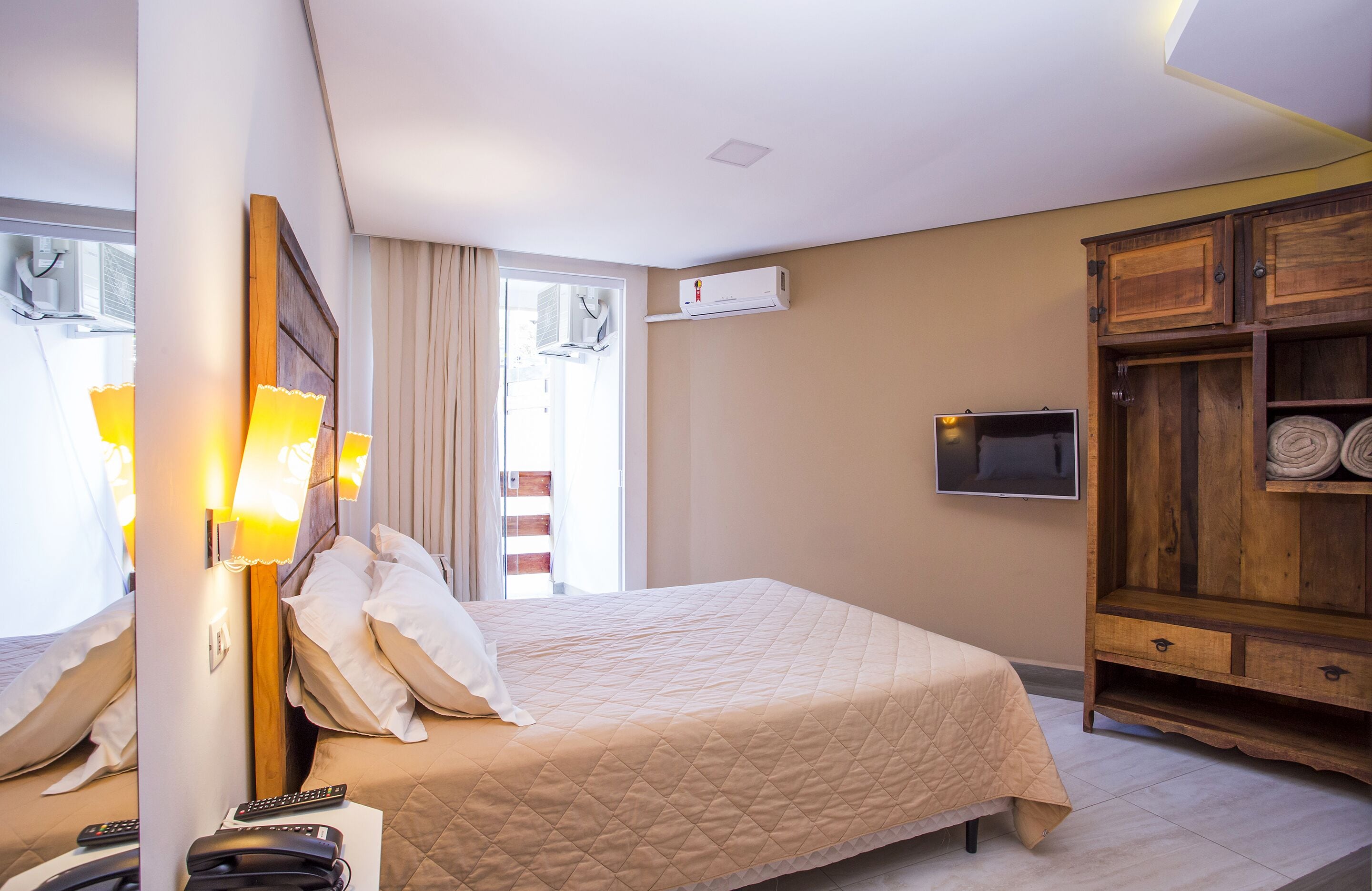 Habitación de hotel con cama, lámparas de noche, televisor, aparador de madera y unidad de aire acondicionado.