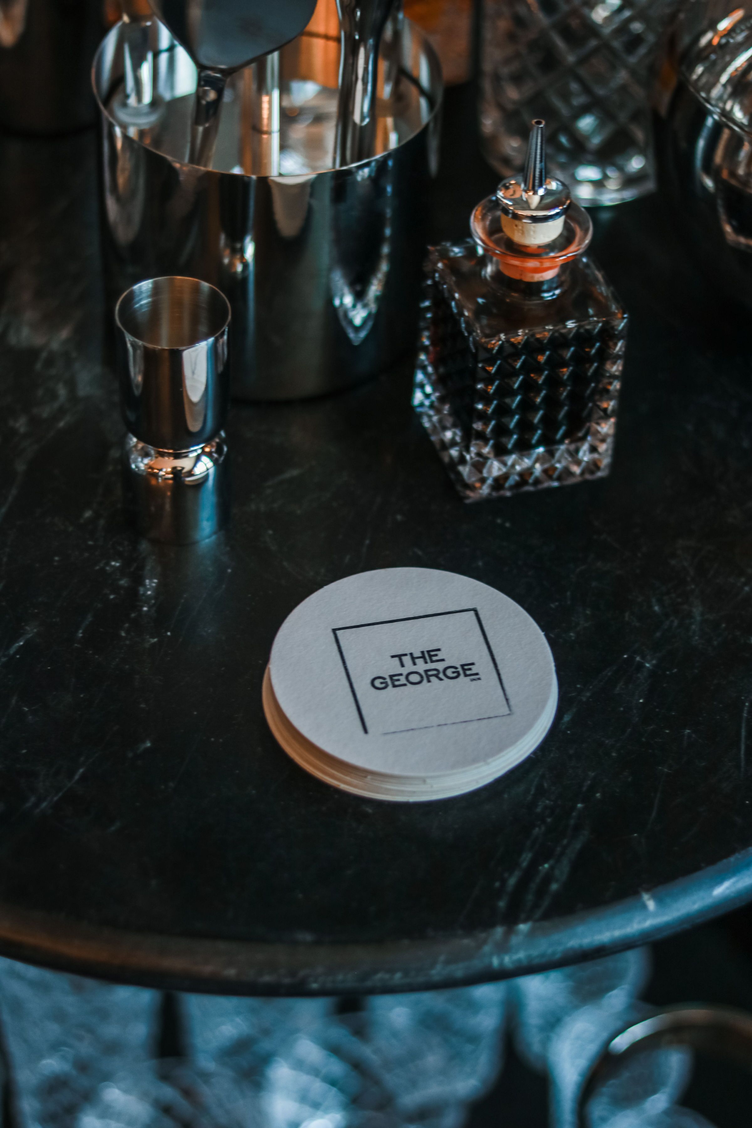 Una zona de bar con un shaker metálico, un vaso pequeño y un posavasos con el texto 'THE GEORGE'.
