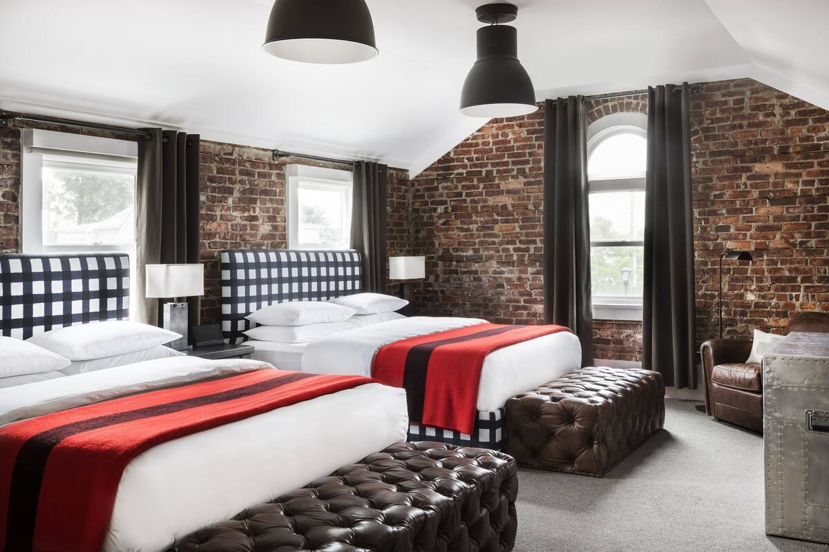 Habitación de hotel con dos camas, un reposapiés de cuero, un sofá de cuero marrón y una pared de ladrillo.