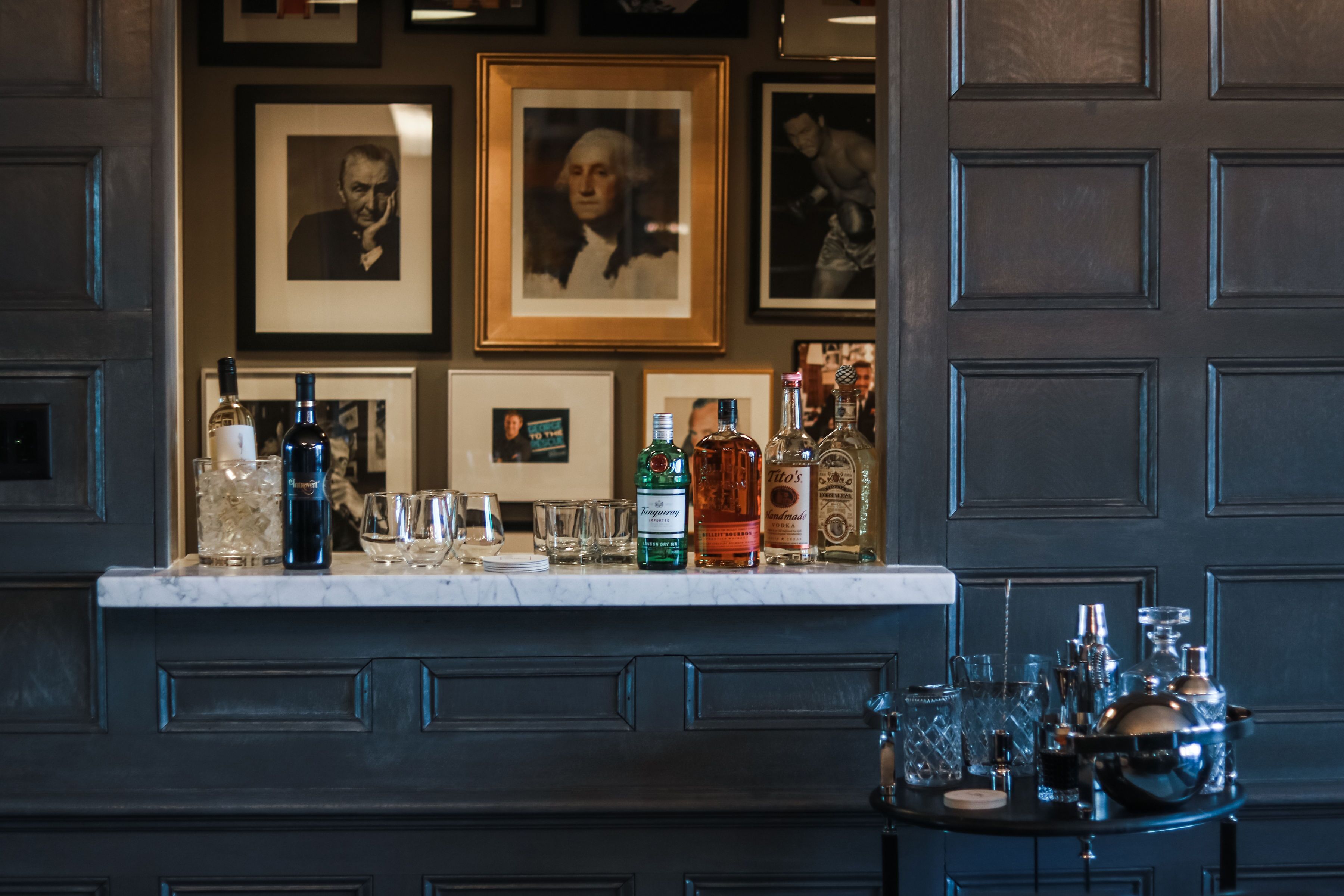 A bar area with a marble countertop, various bottles, glasses, and framed pictures on the wall.