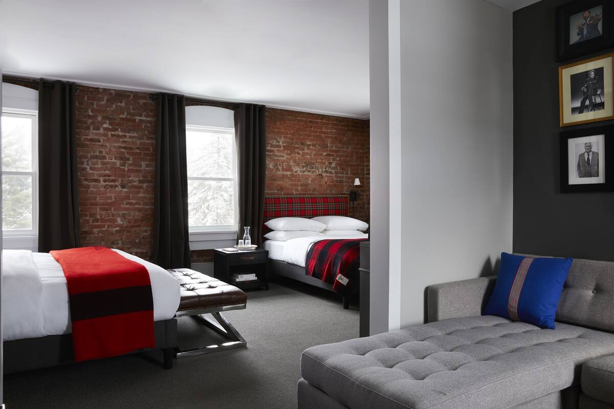 Una habitación de hotel moderna con una pared de ladrillo decorativa, una cama con cabecera de cuadros rojos, un sofá gris y una mesita de noche con lámpara.