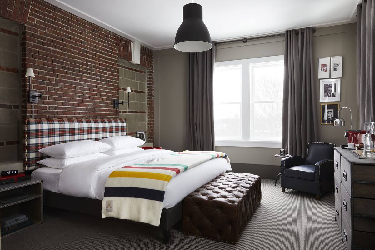 Habitación de hotel con pared de ladrillo, una cama grande con cabecero de cuadros, un escritorio con silla y una ventana con cortinas.