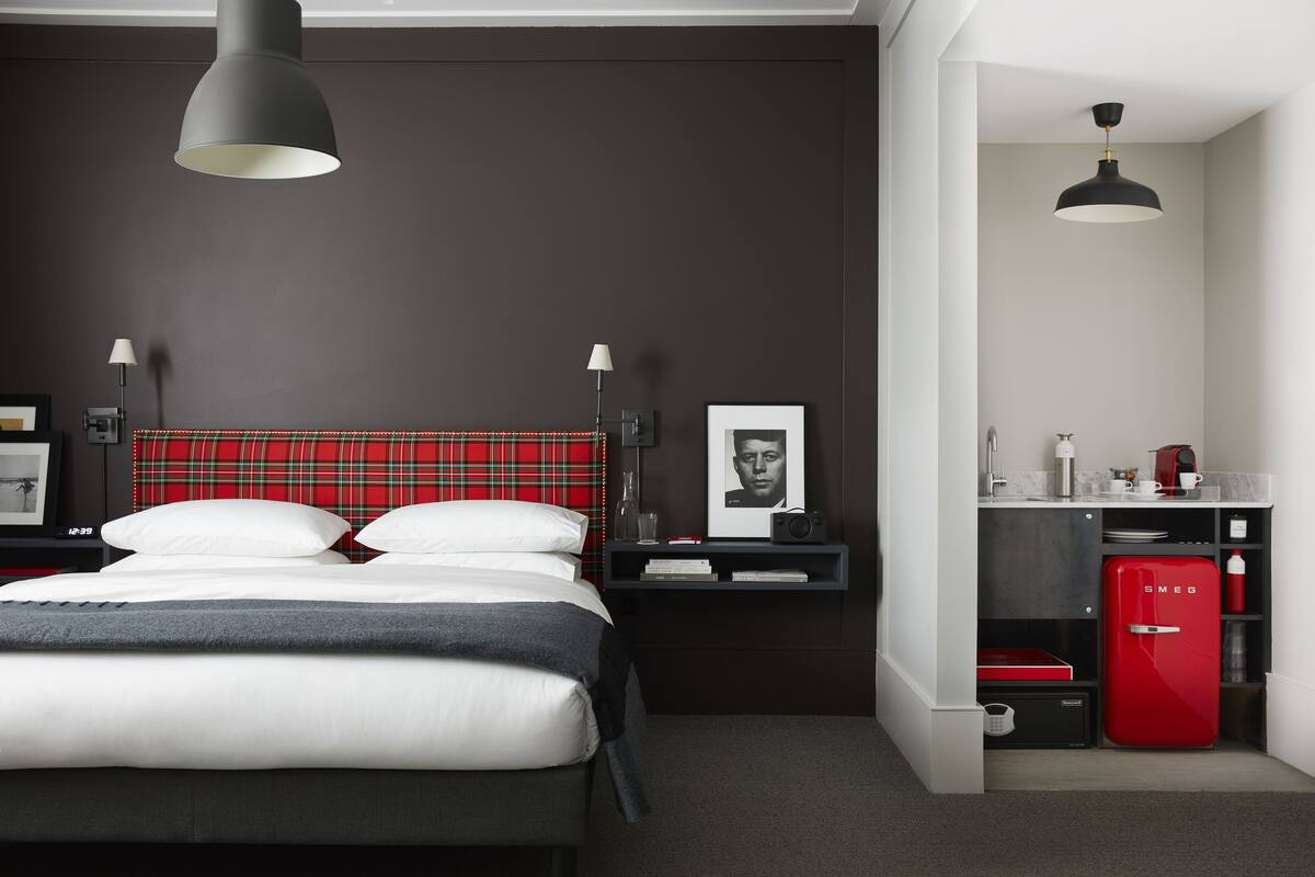 Un dormitorio moderno con cama, mesitas de noche, un refrigerador rojo y una pequeña kitchenette.