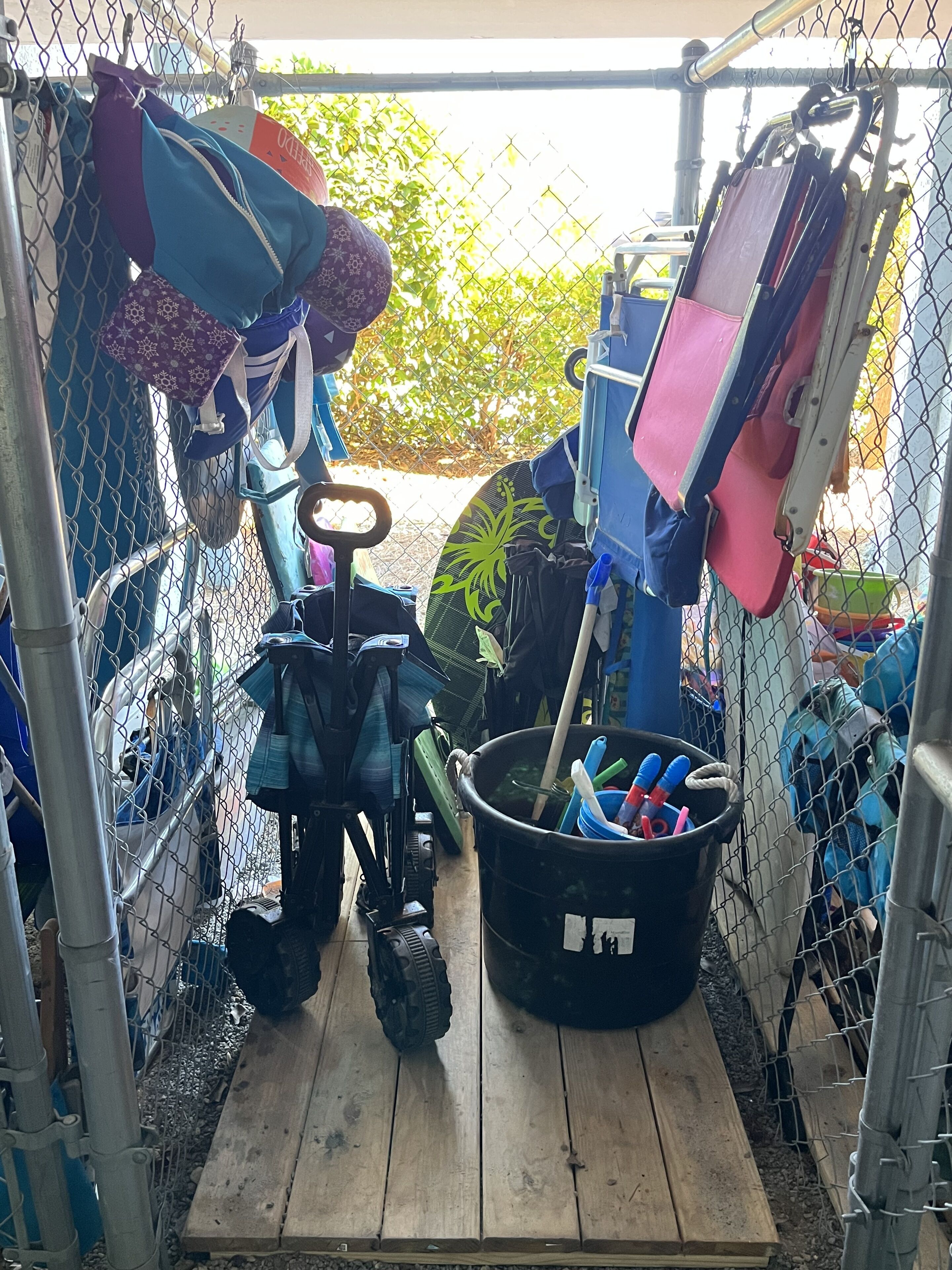 A narrow outdoor storage area with a wooden bench, a black bucket with cleaning supplies, and various bags and items hanging on a chain-link fence.