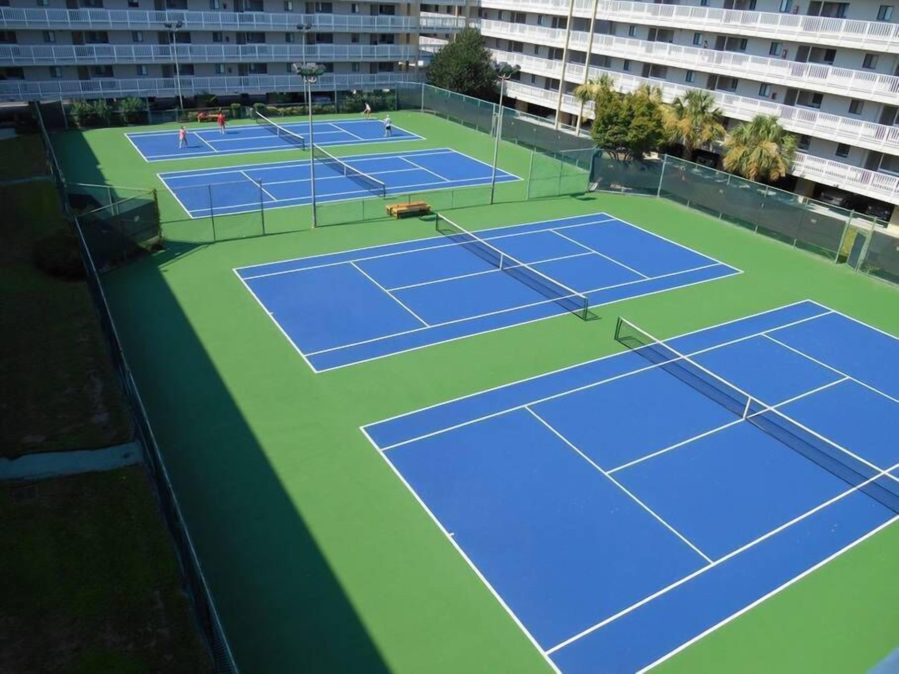 An outdoor tennis court with multiple courts, surrounded by a fence and a multi-story building in the background.