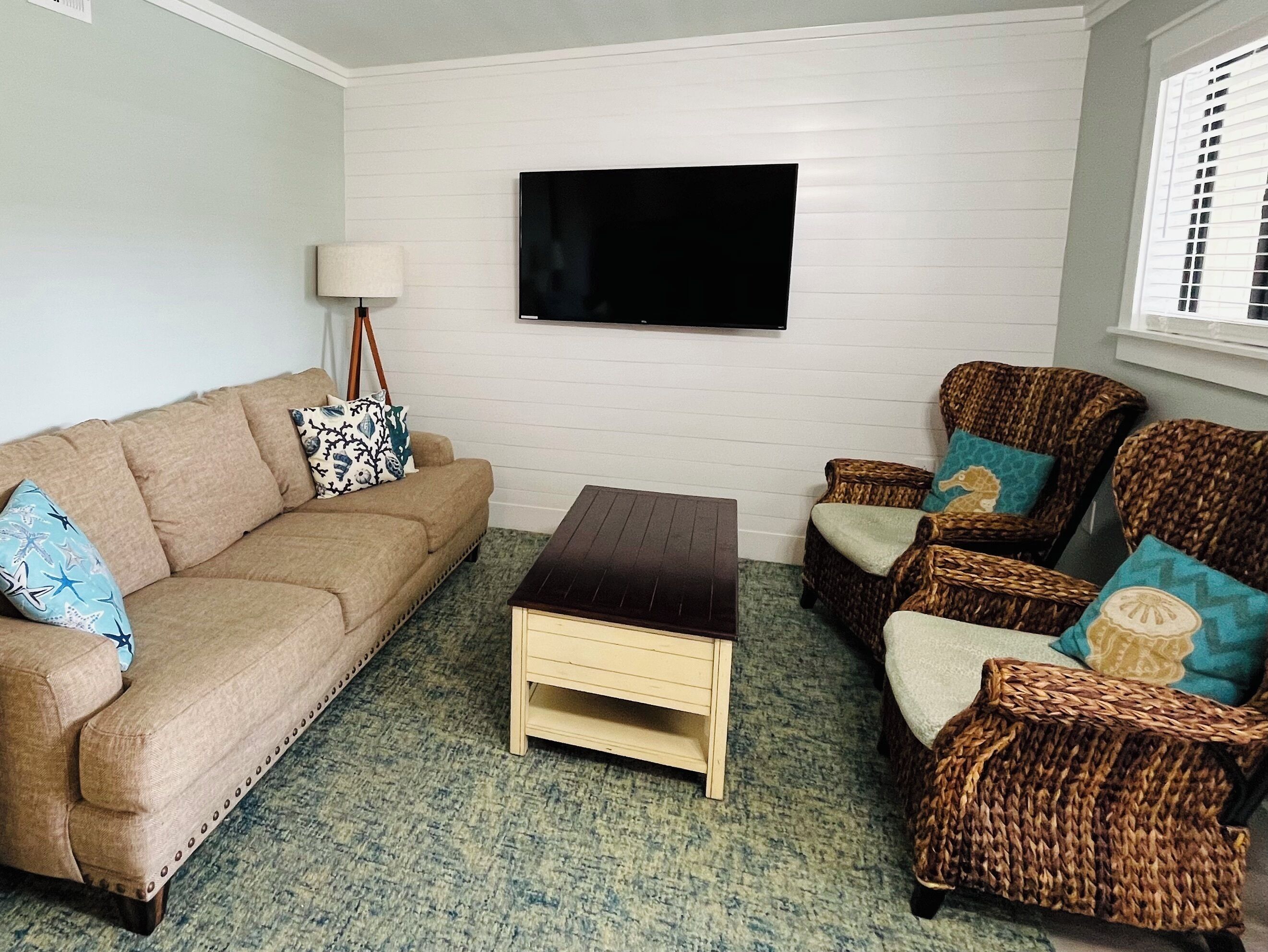 A living room with a beige sofa, a dark wooden coffee table, wicker chairs, and a flat-screen TV mounted on the wall.