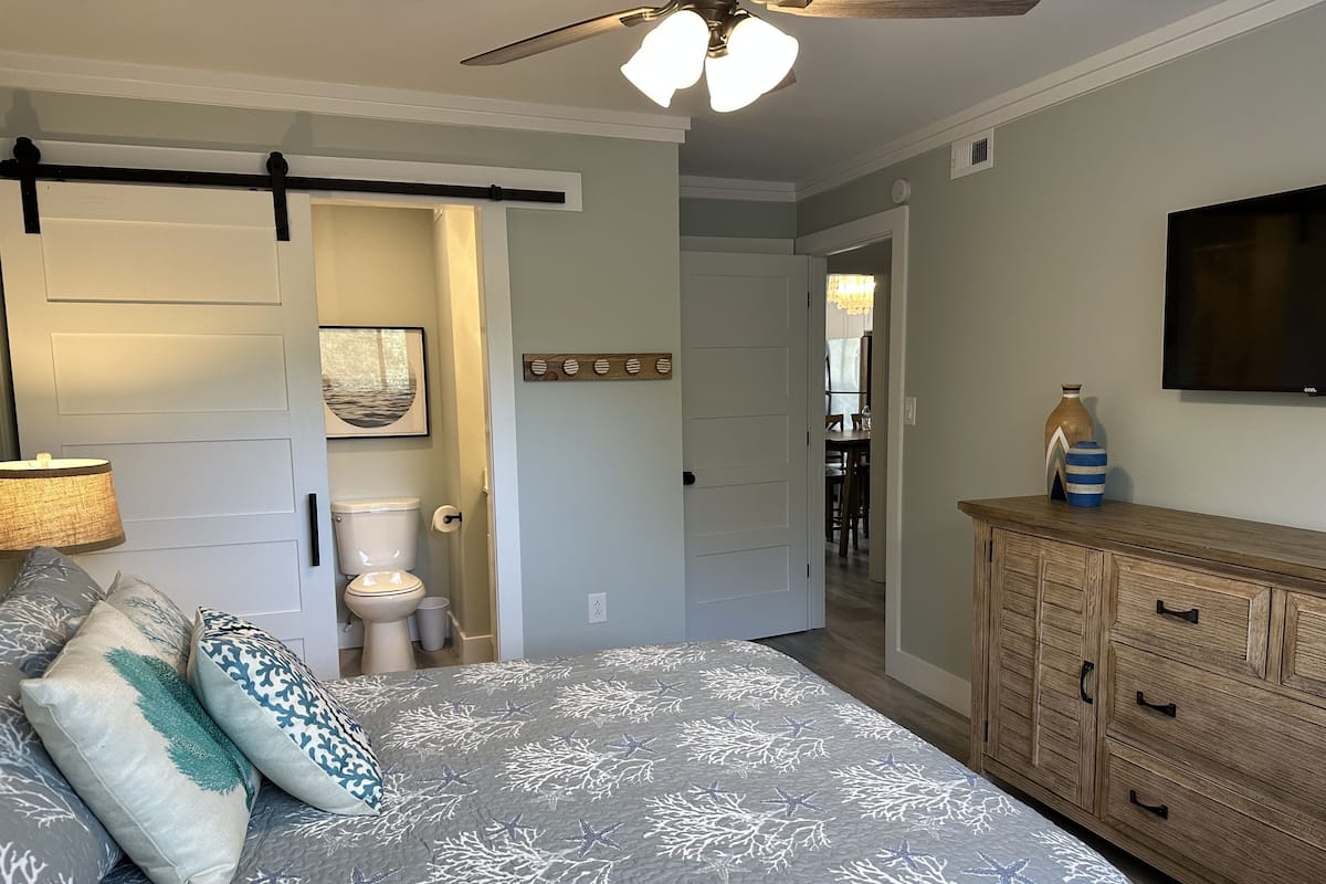 A bedroom with a bed, a wooden dresser, a ceiling fan, and a TV on the wall.