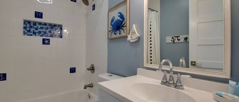 Une salle de bain avec une baignoire blanche, un lavabo et une douche. Le mur est décoré de carreaux et un tableau encadré est accroché au-dessus du lavabo.