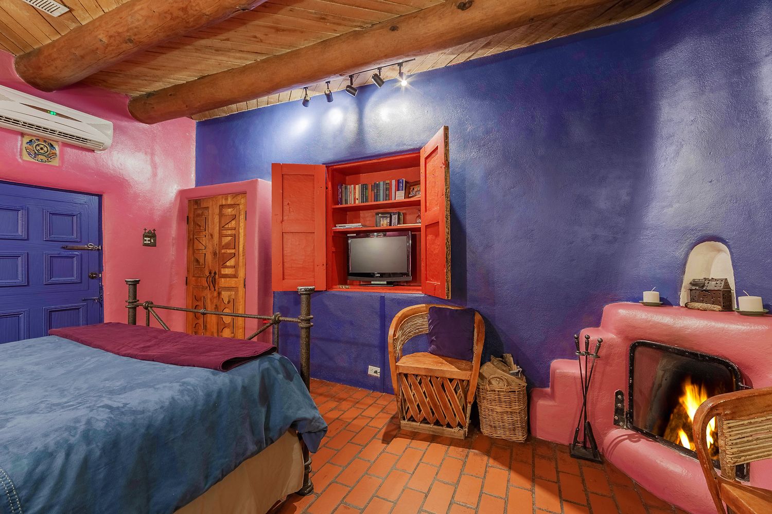 A room with a bed, a television, a bookshelf, a chair, a fireplace, and a red door.