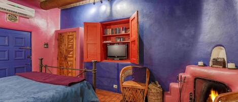 A room with a bed, a television, a bookshelf, a chair, a fireplace, and a red door.