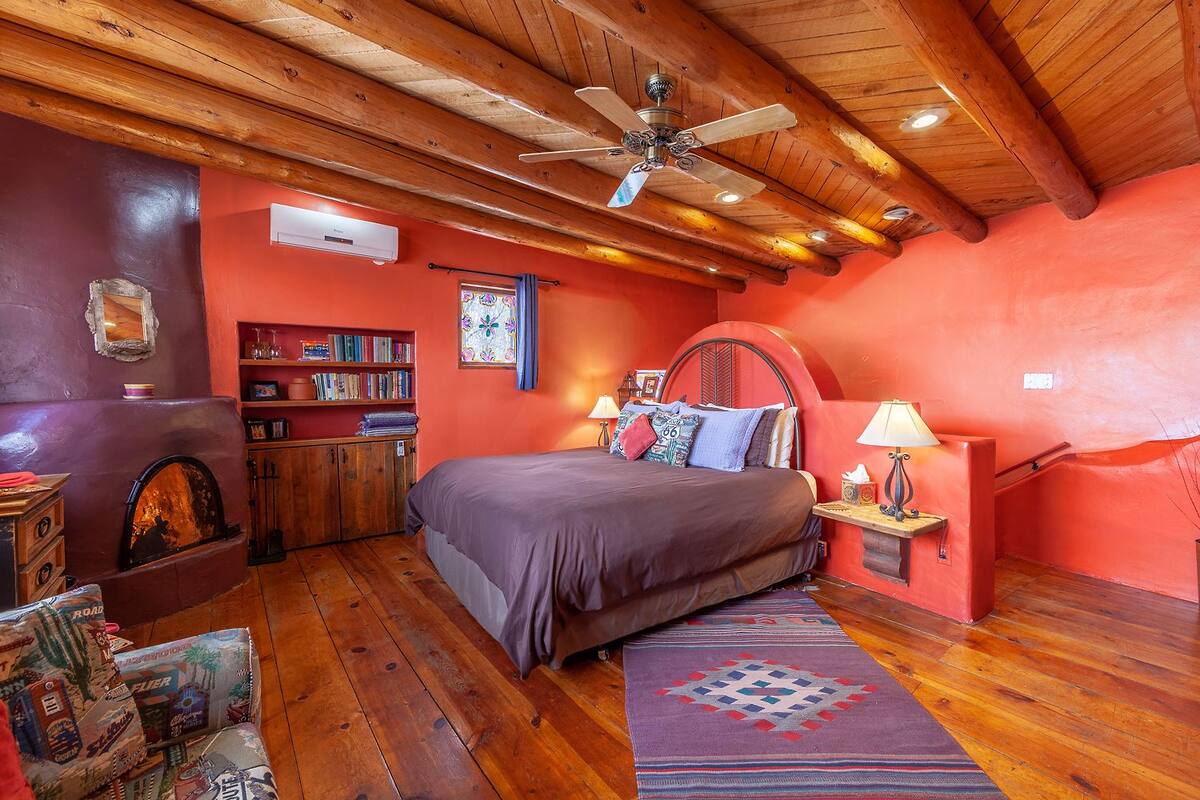 A cozy bedroom with a wooden ceiling, a bed with a headboard, a fireplace, a bookshelf, and a window with a view.