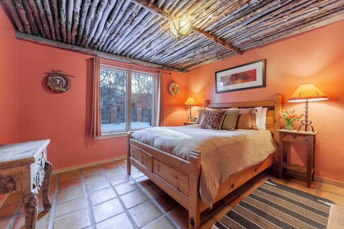 A bedroom with a wooden bed, two bedside tables with lamps, a window with curtains, and a rustic ceiling.