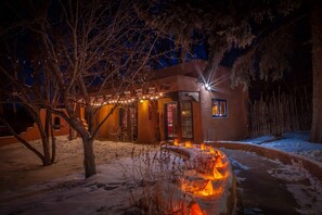 A rustic building with a snow-covered ground, a fire pit with burning logs, and string lights.