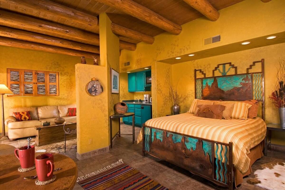 A cozy bedroom with a wooden ceiling, a bed with a unique headboard, a small kitchenette, and a dining area with a table and chairs.