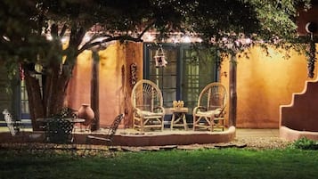 An outdoor seating area with wicker chairs and a table, surrounded by trees and a building with a warm, yellowish wall.