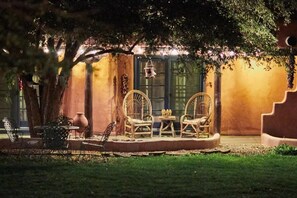 An outdoor seating area with wicker chairs and a table, surrounded by trees and a building with a warm, yellowish wall.