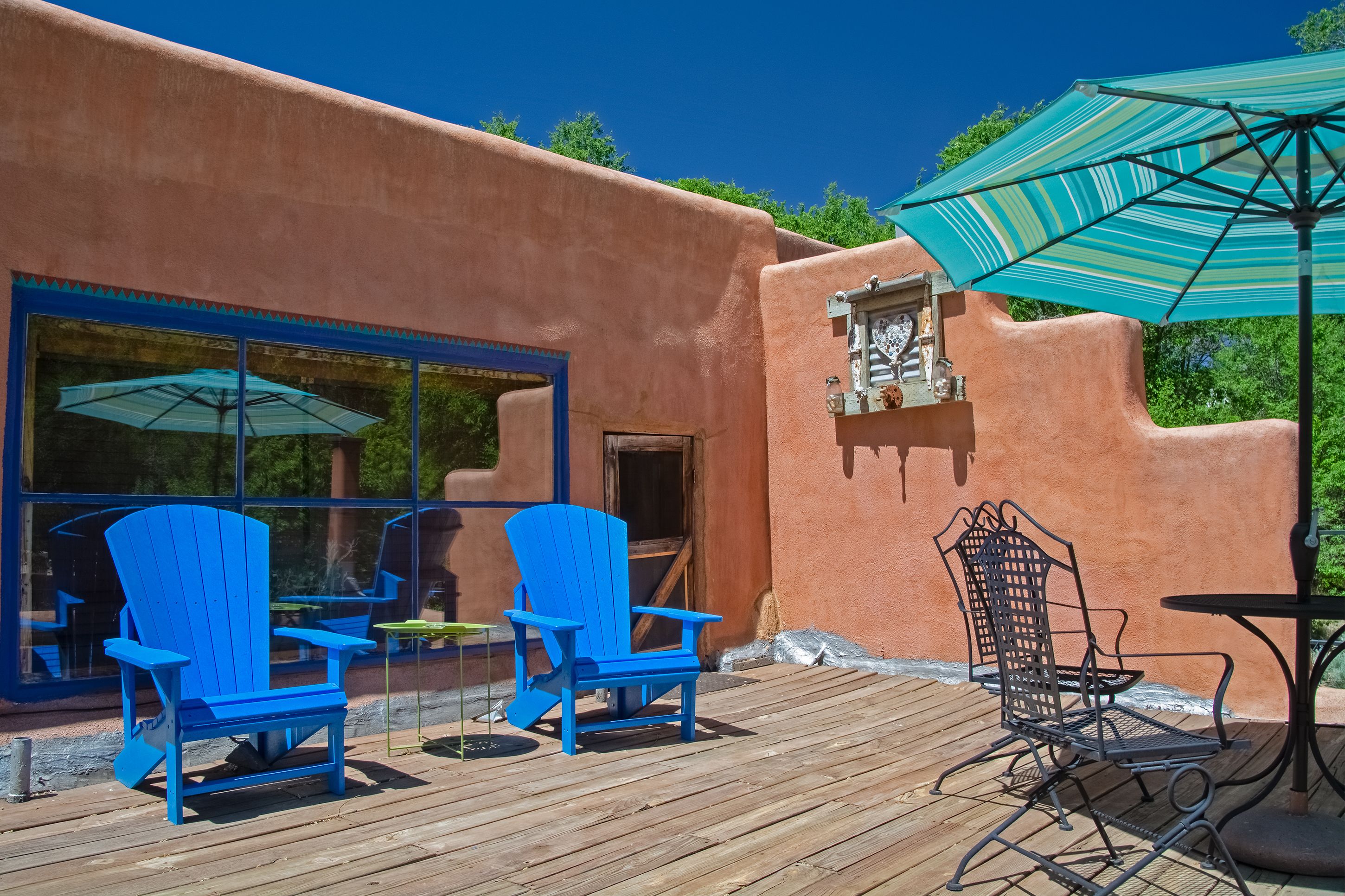Una terraza de madera con sillas Adirondack azules y una silla de metal, un paraguas turquesa, y un edificio rústico con una ventana y una puerta.
