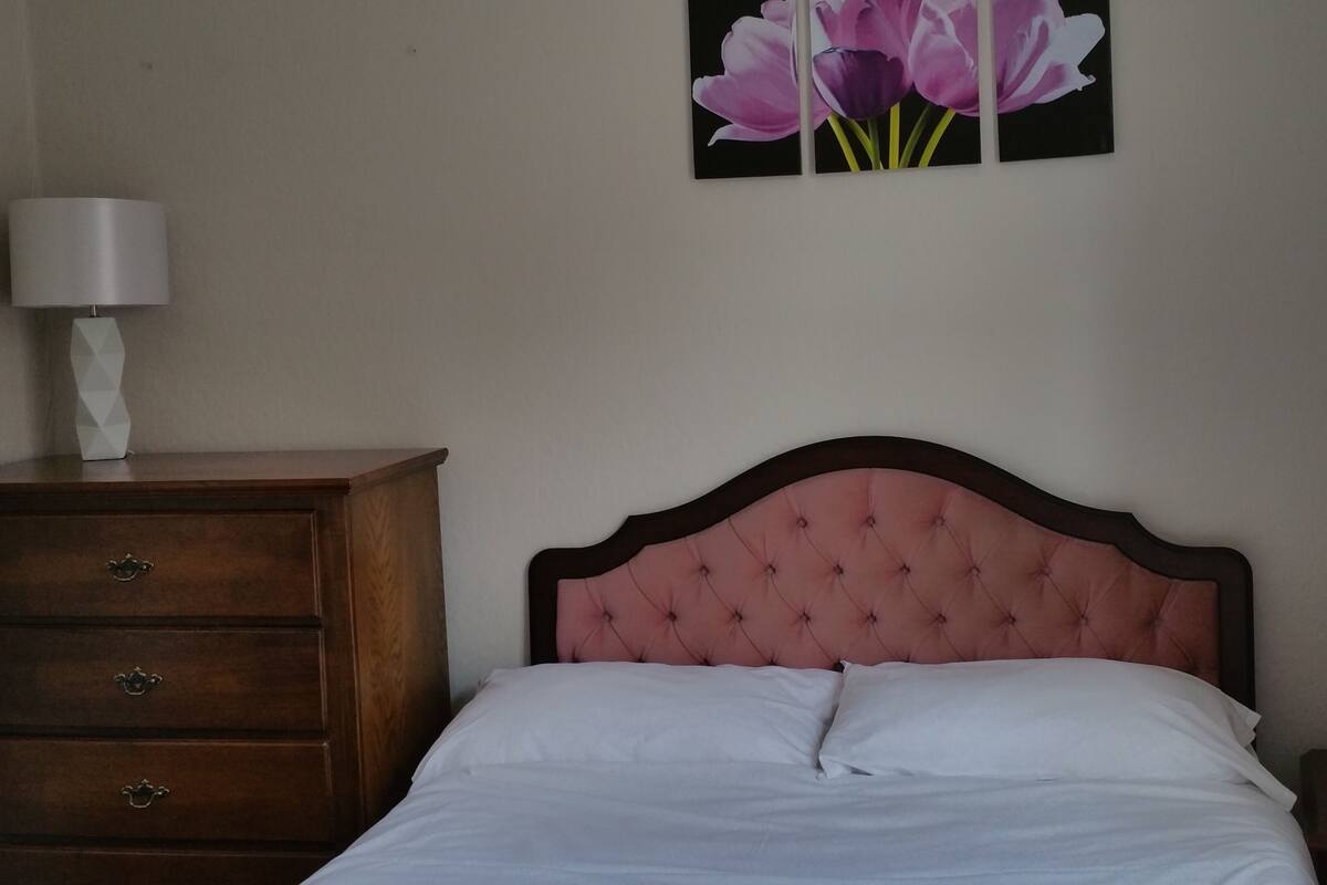 A bedroom with a bed, a wooden dresser, a nightstand, a lamp, and a wall-mounted painting of flowers.