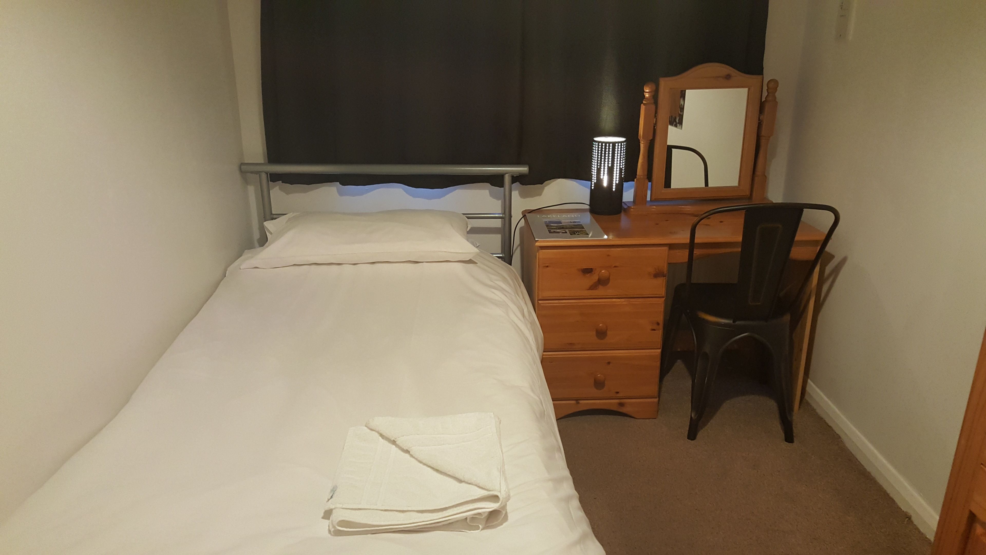 Een kleine kamer met een enkel bed, een houten dressoir, een stoel en een bureau.