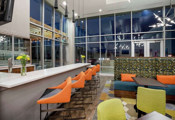 A modern bar area with a long counter, multiple orange bar stools, and a seating area with a sofa and chairs.