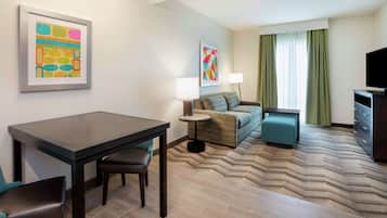 Een hotelkamer met een eettafel, twee stoelen, een bank, een bijzettafeltje met een lamp, een televisie en een kamerbreed tapijt met een patroon.