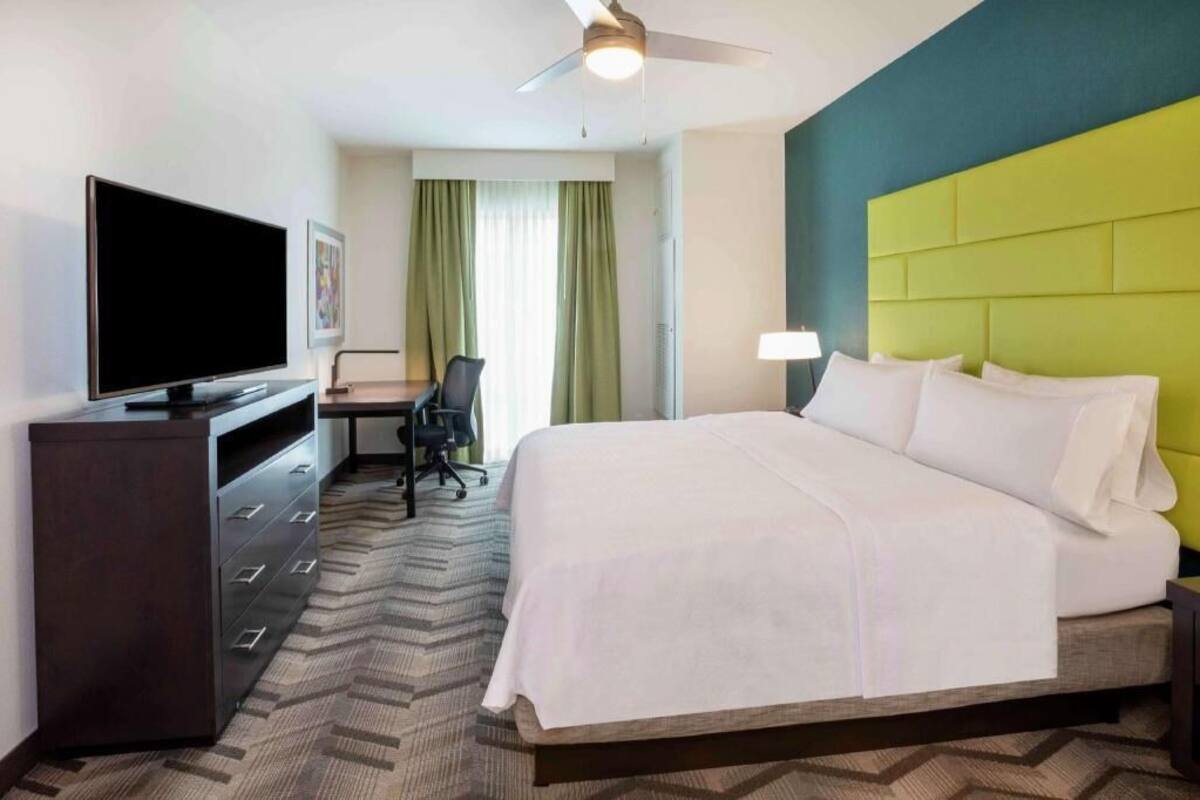 A hotel room with a bed, a desk with a chair, a television, and a ceiling fan.