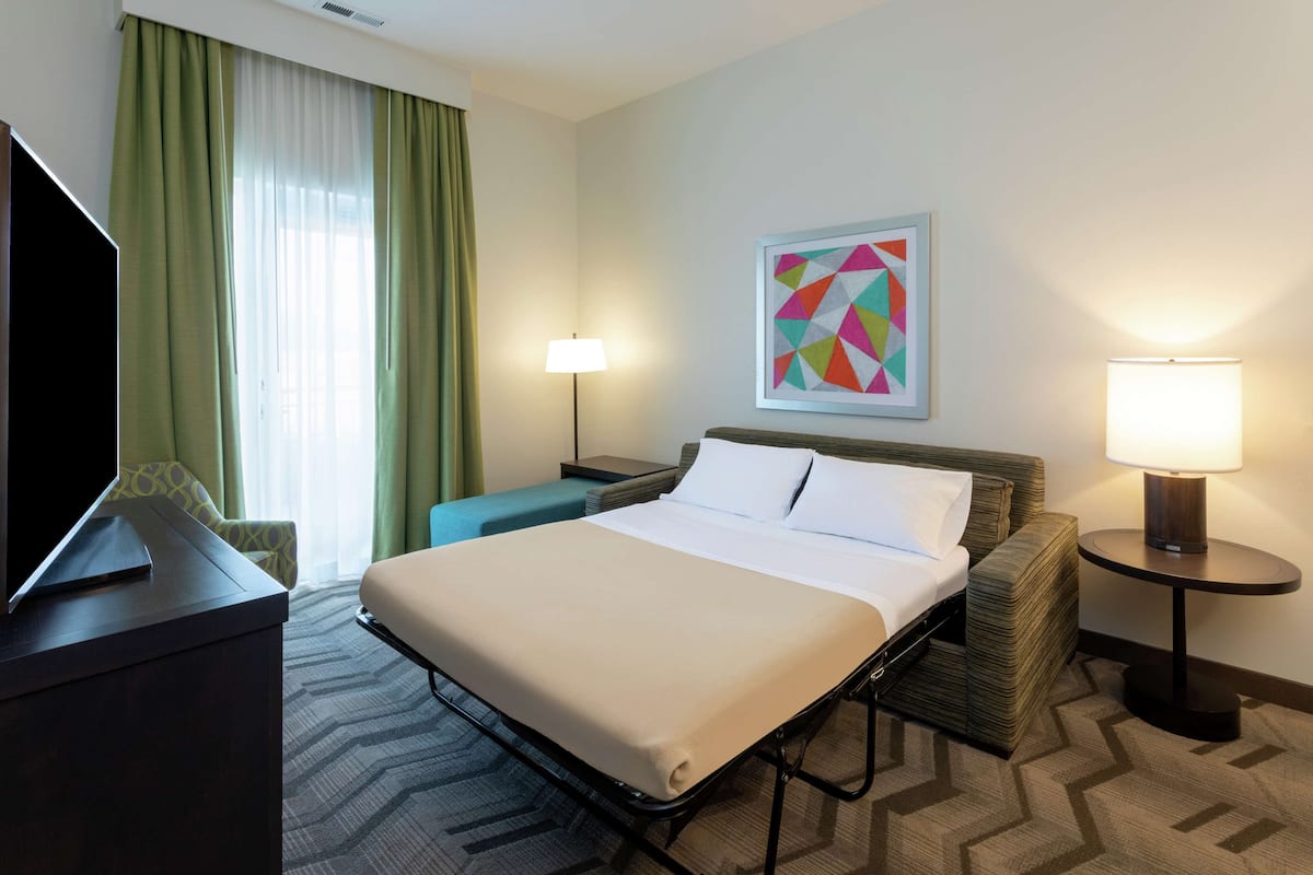 A hotel room with a bed, a TV, a desk, a lamp, and a painting on the wall.
