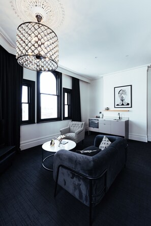 A modern living room with a black and white sofa, a round coffee table, and a pendant light. There is a window with black curtains, a white wall with a framed artwork, and a small kitchenette area.