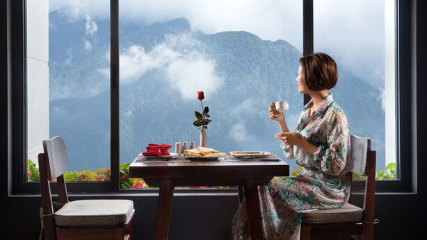 A person seated at a table with a view of mountains through a large window. - The View Sapa Hotel (Sa Pa)