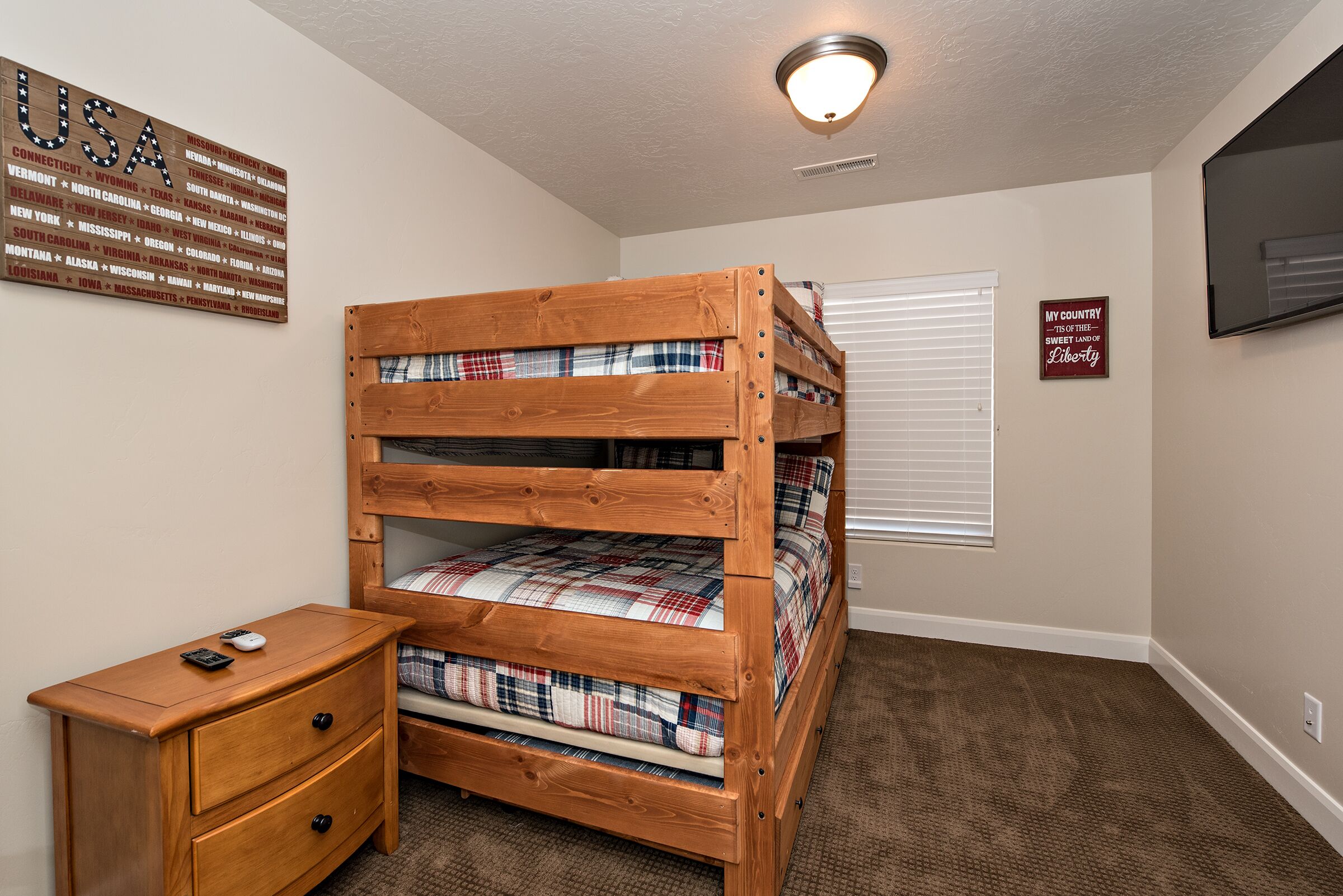A bunk bed with plaid bedding, a wooden nightstand, a wall map of the USA, and a sign stating "My Country, the Sweet Land of Liberty."