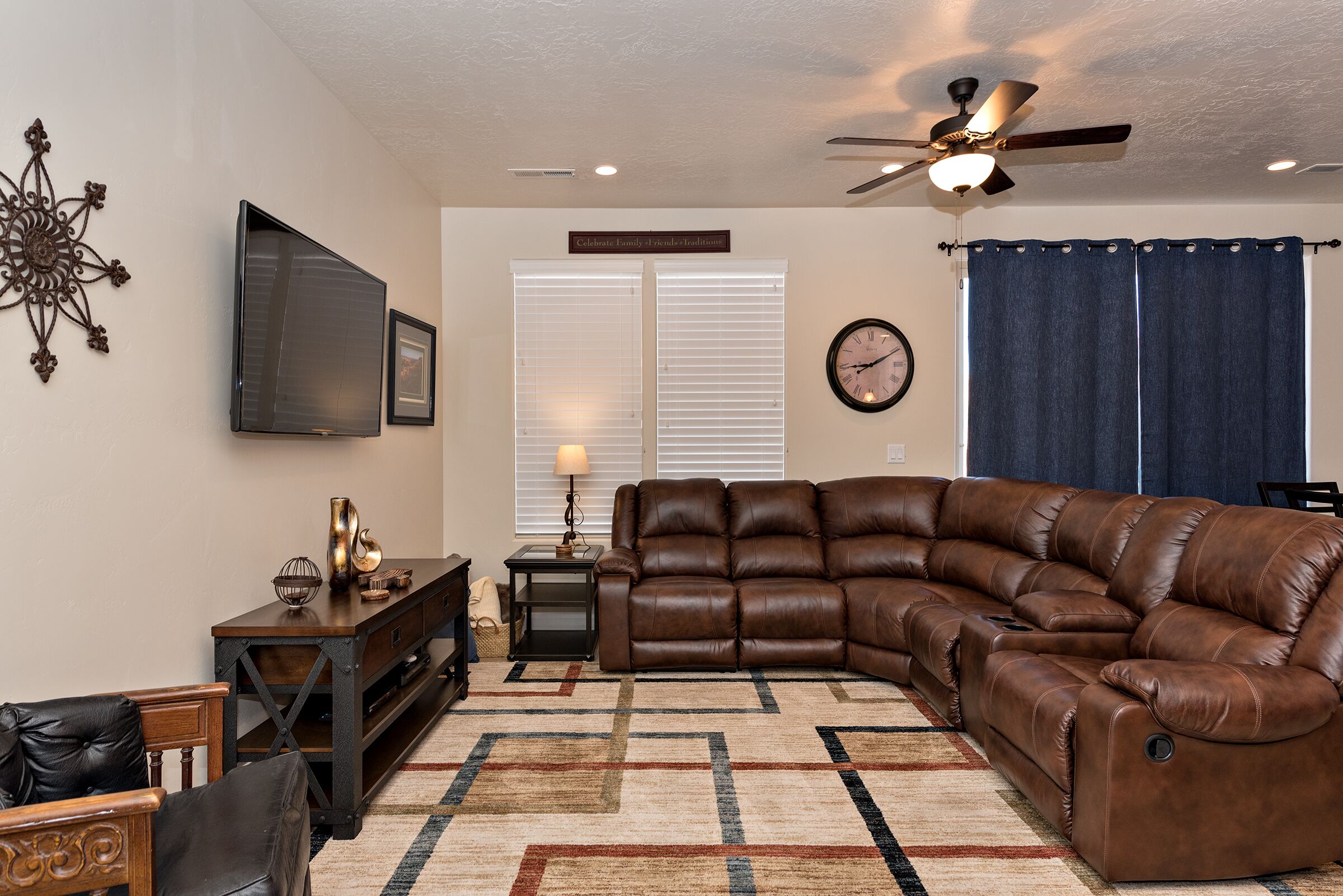 A living room with a sectional sofa, a television mounted on the wall, a ceiling fan, and a decorative wall piece.