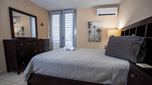 A bedroom with a bed, a dresser, a mirror, a painting, and an air conditioner.