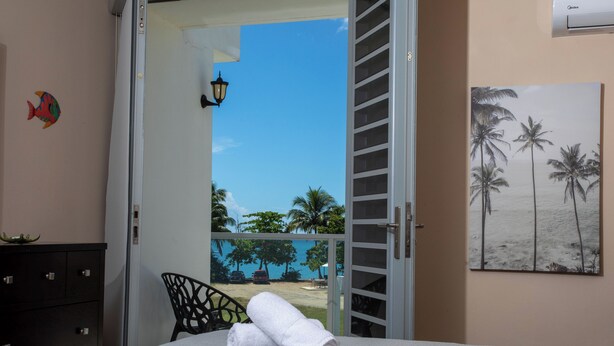 A room with a bed, a chair, a door leading to a balcony with a view of palm trees and the sea.