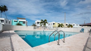 A swimming pool with a metal ladder, surrounded by a white fence and palm trees, with buildings in the background.