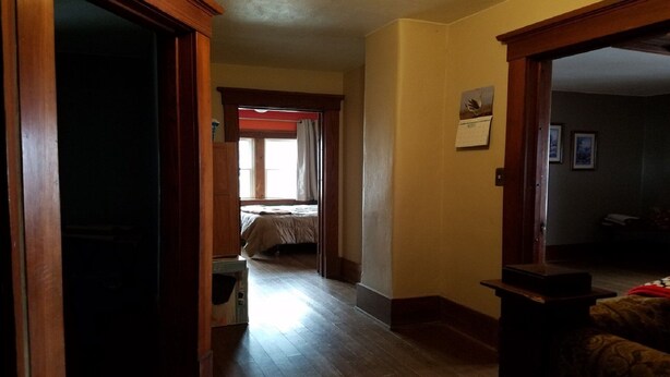 A narrow hallway with wooden flooring leading to a bedroom with a bed and a window.