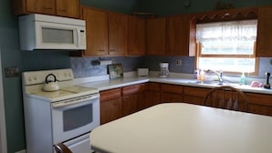 A kitchen with wooden cabinets, a white stove, a microwave, a window with lace curtains, and a table.