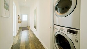 Un cuarto de lavado con dos lavadoras de carga frontal apiladas, piso de madera y una puerta al final del pasillo.