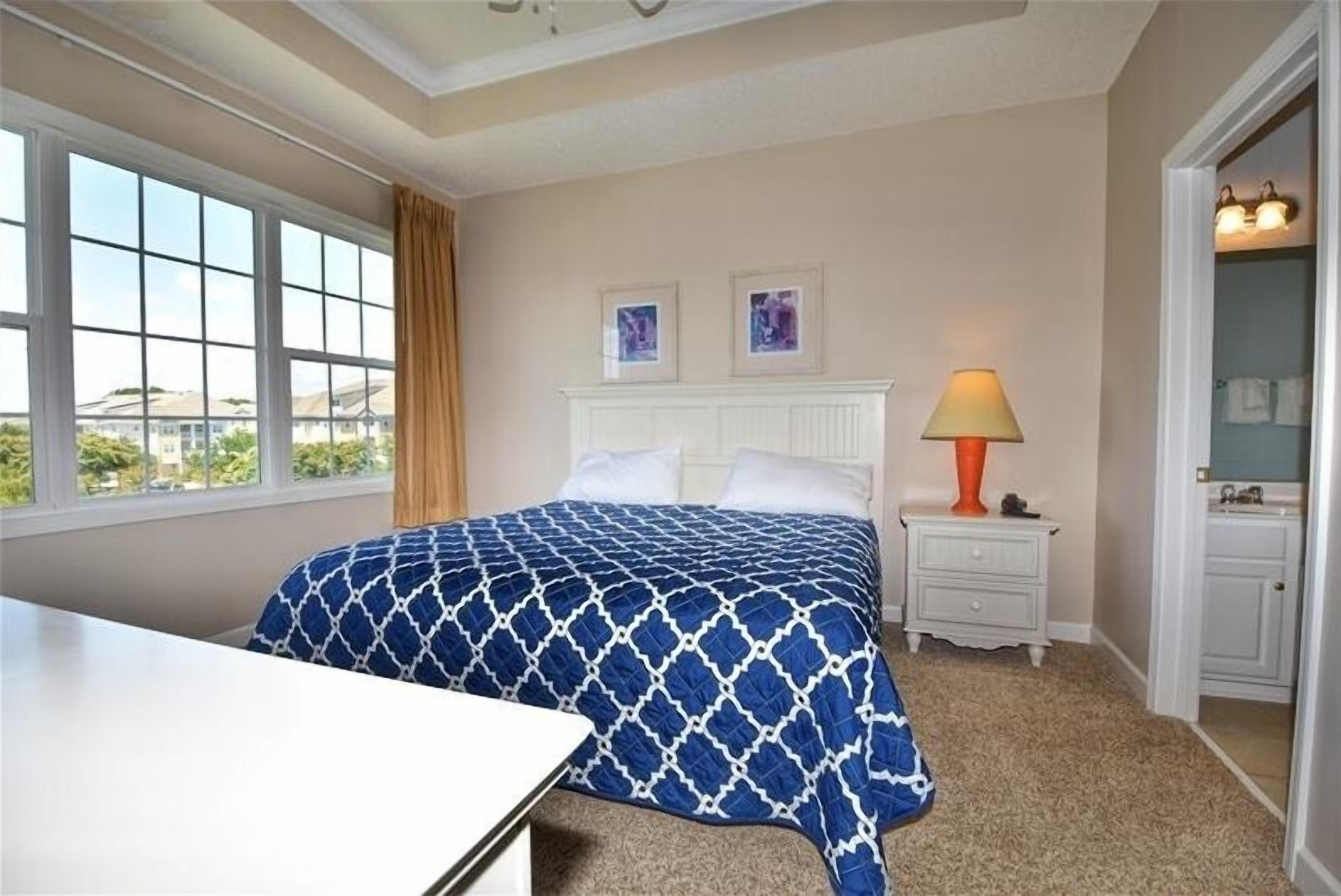 A bedroom with a large window, a bed with a patterned bedspread, a bedside table with a lamp, and a bathroom visible through an open door.