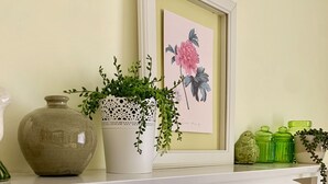 Una repisa blanca con una obra de arte floral enmarco, una planta en maceta, frascos de vidrio verde y un espejo decorativo.