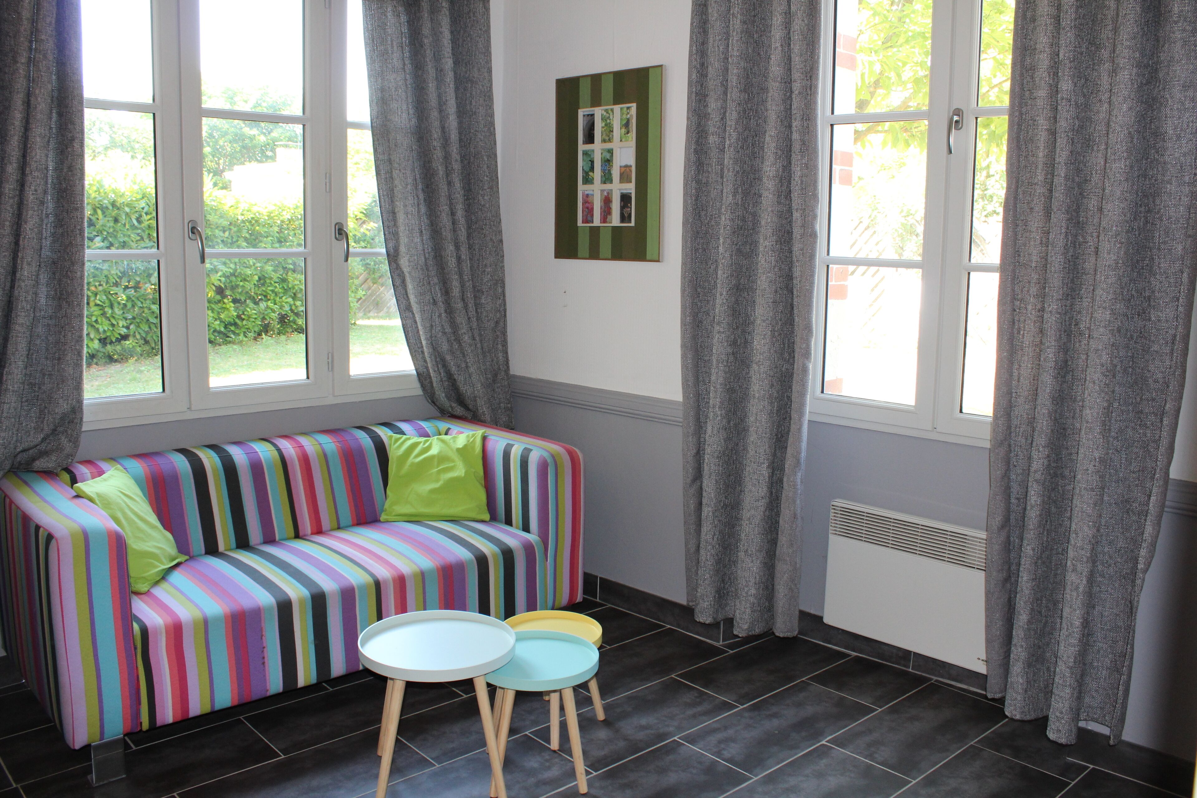 Een kamer met een kleurrijke gestreepte bank, een kleine tafel en twee stoelen. Gordijnen hangen voor de ramen, en er is een radiator op de muur te zien.