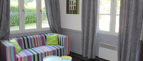 A room with a colorful striped sofa, a small table, and two chairs. There are curtains on the windows, and a radiator is visible on the wall.